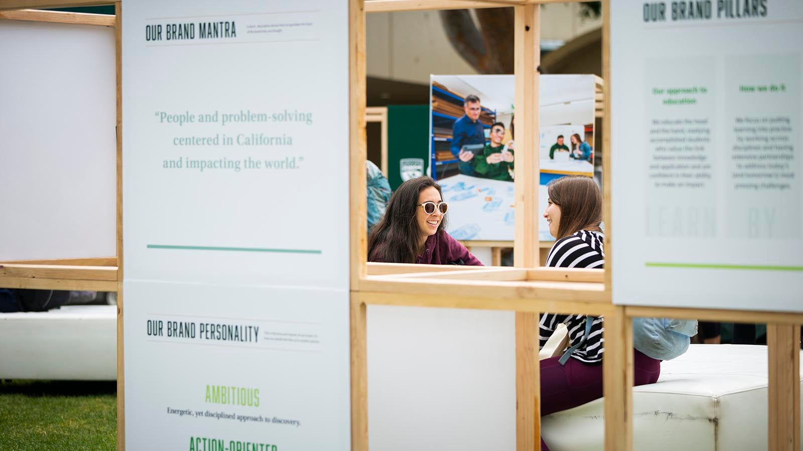 Foreground is a wooden display showing brand information with two students sitting on a cushioned bench in the background.
