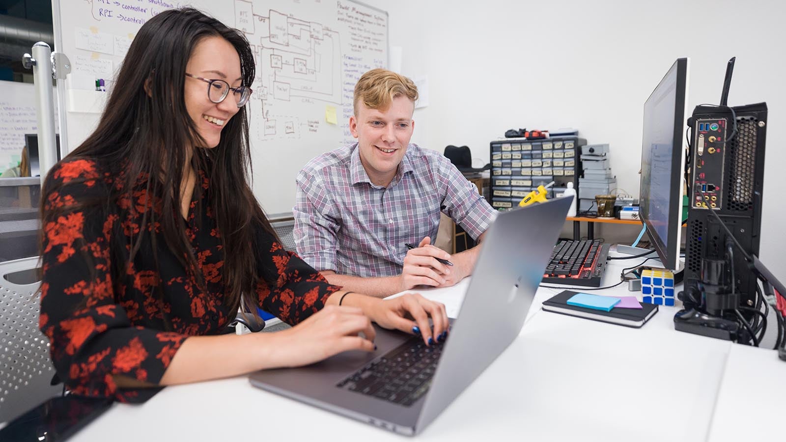 Two students chat while seated at a table with a laptop open in front of them. 