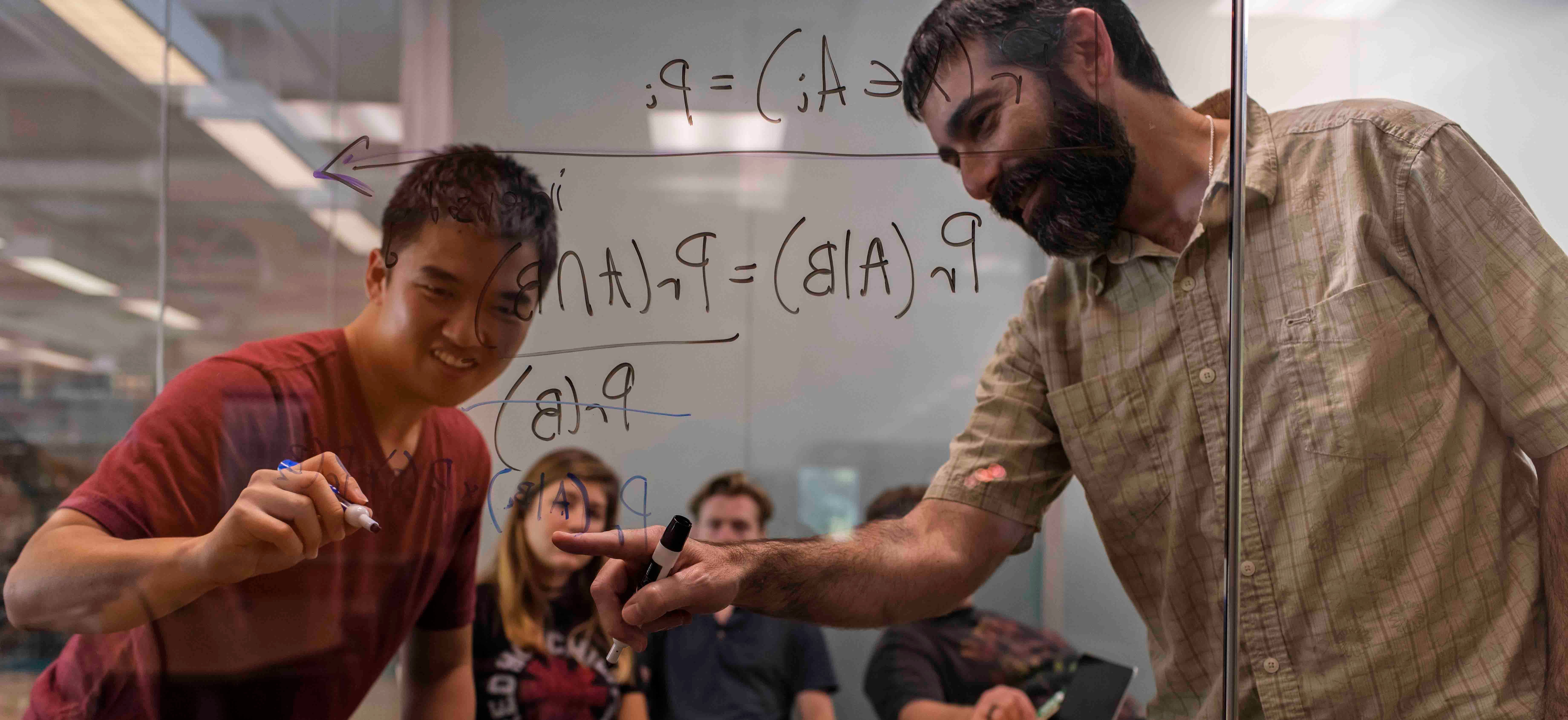 Statistics Professor Andrew Schaffner works with a student writing equations on glass walls