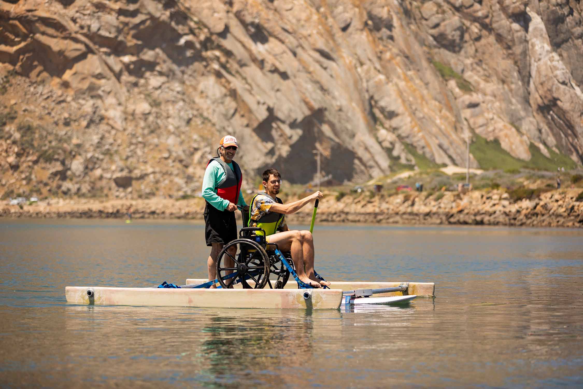 Adapted Paddling Program member helps a participant in a wheelchair-adapted stand-up paddle board with Morro Rock in the background