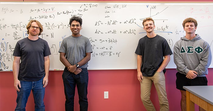 Assistant Professor Ben Shlaer with students stand before a whiteboard with cosmology-related formulas and calculations