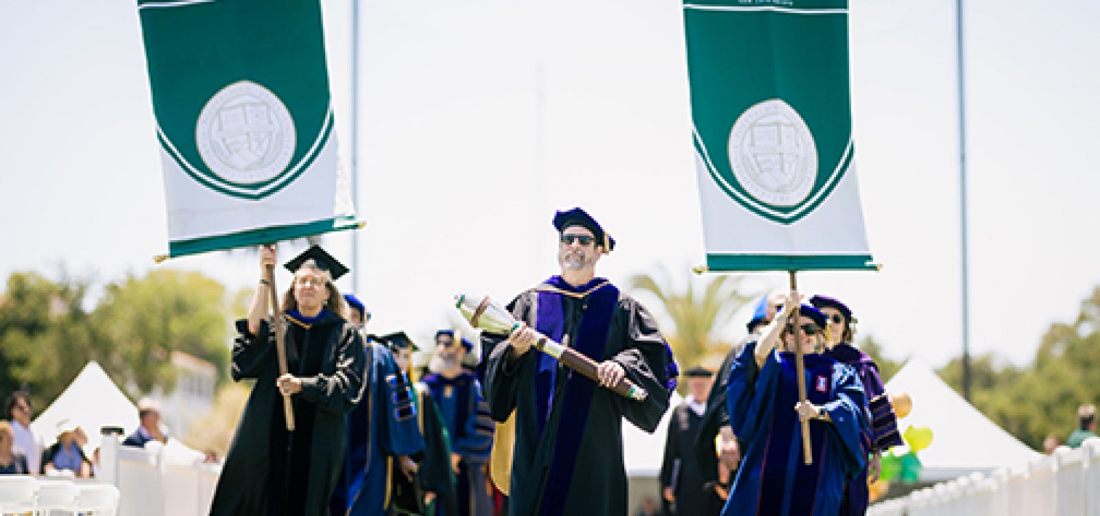 Commencement stage officials holding college banner including a man carrying the ceremonial mace enter the stadium for the 2023 Cal Poly commencement