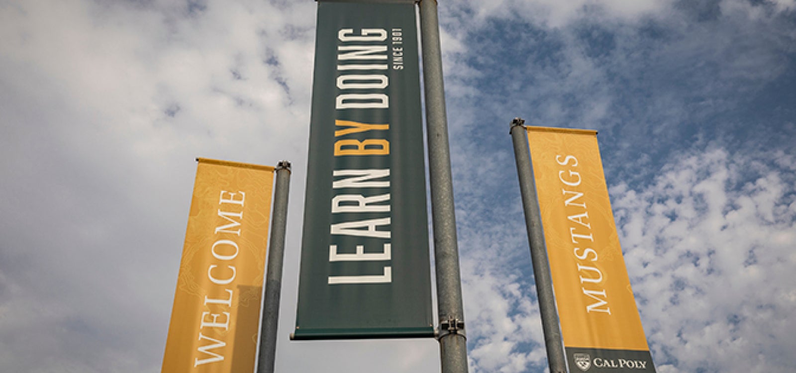 A trio of campus entrance banners say Welcome, Mustangs, and Learn by Doing -- Since 1901