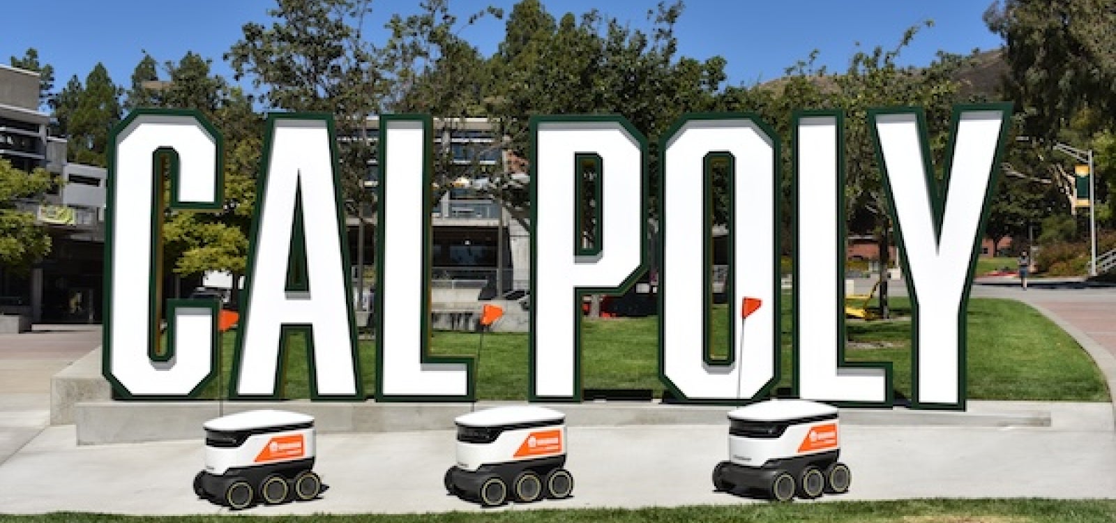 Three Starship robots, which are six-wheeled carriers under 30 inches long, 2 feet wide and tall, with a small orange flag and Grubhub branding, are parked in front of a structure of the letters spelling out Cal Poly in the University Union on campus.