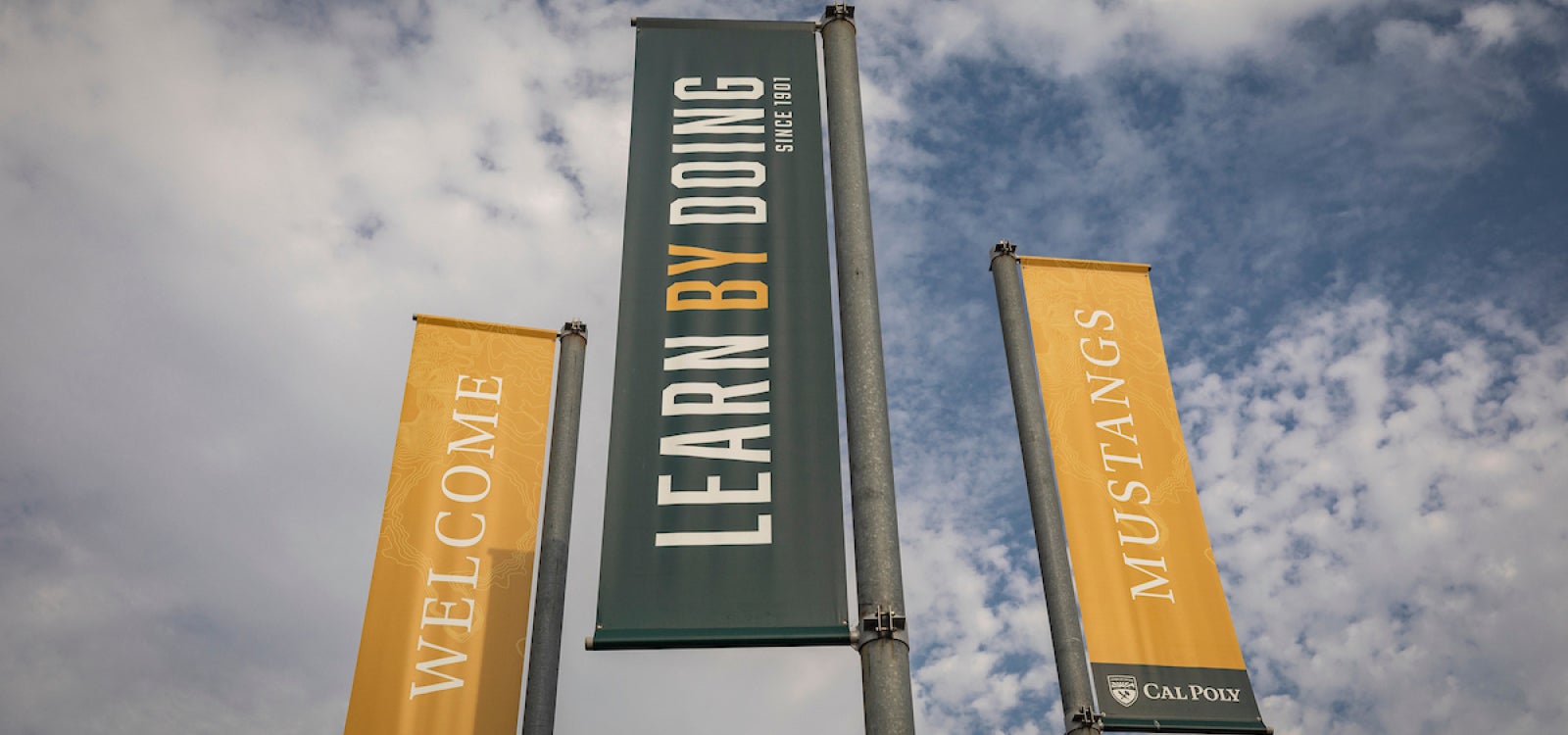 A trio of Cal Poly welcoming banners under a partly cloudy sky