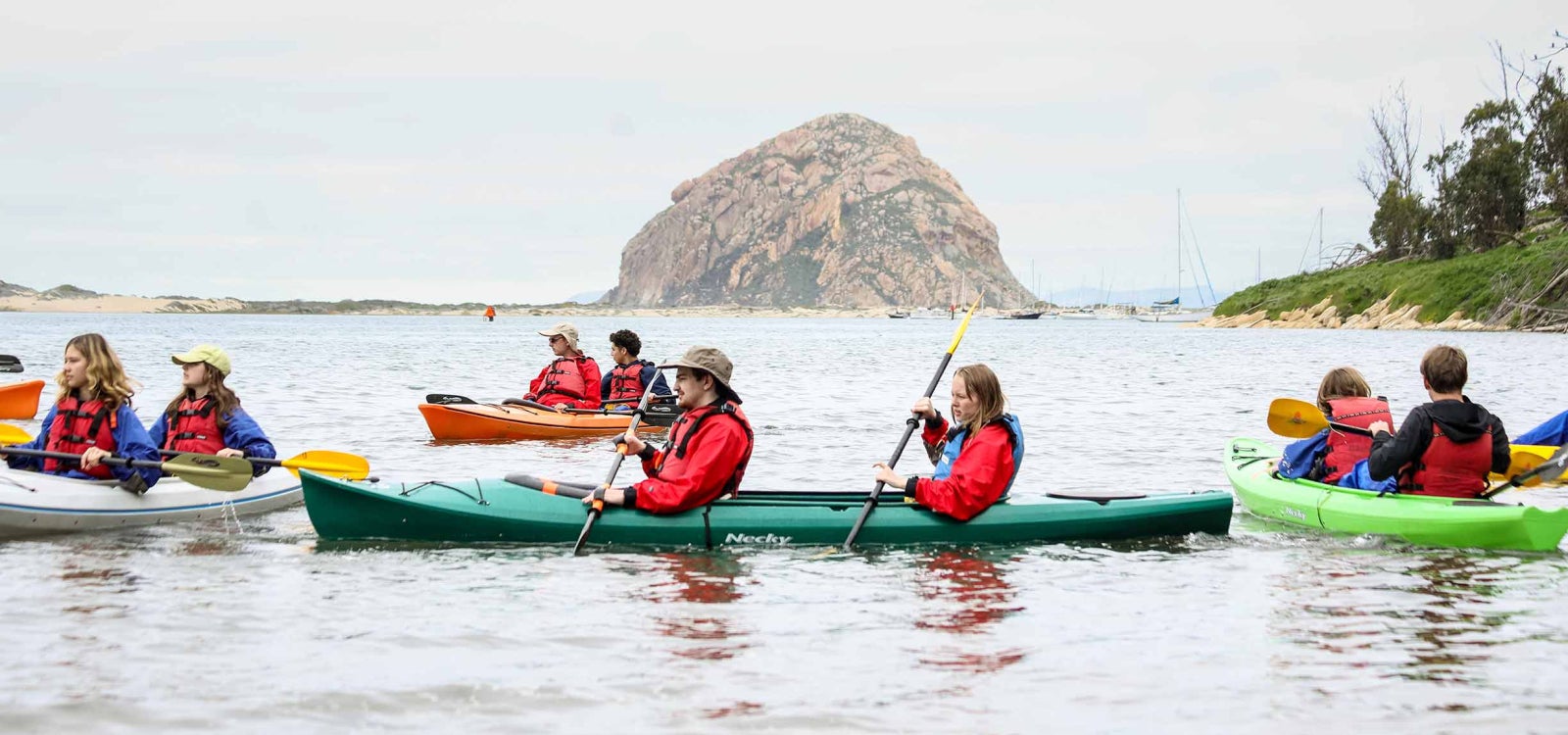 Groups of tudents and their clients paddle kayaks in Morro Bay with Morro Rock in the background