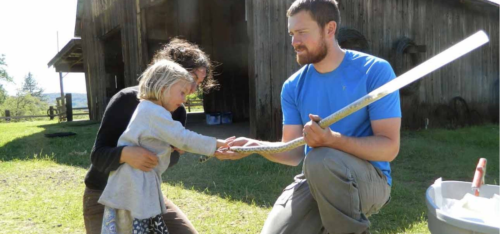 A child held by her mother examines the tail of a snake that is in a protective tube held by a man