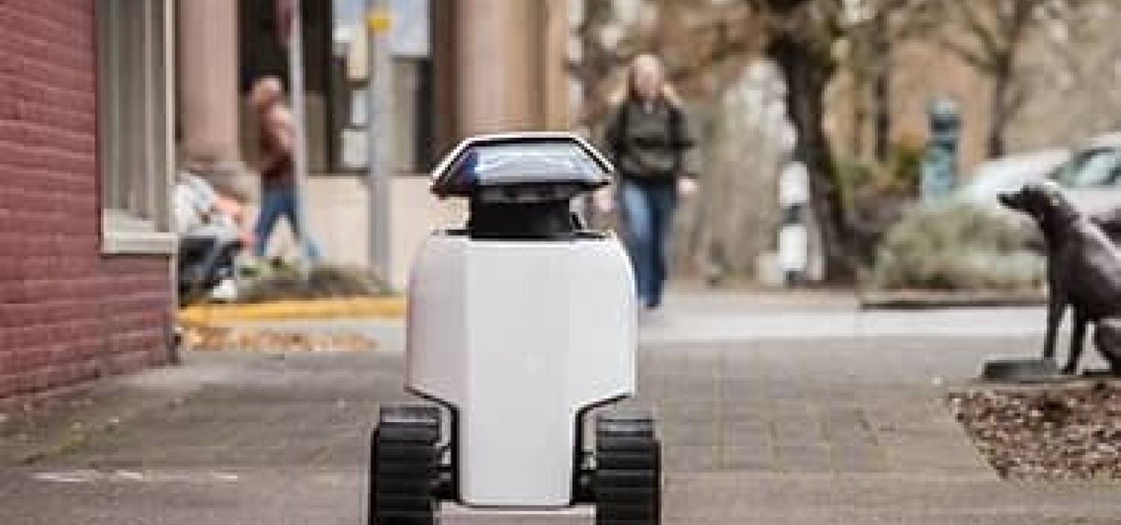 A Daxbot robot rolls along a sidewalk navigating past buildings and a street bench