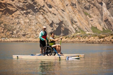 Adapted Paddling Program member helps a participant in a wheelchair-adapted stand-up paddle board with Morro Rock in the background