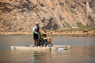 Adapted Paddling Program member helps a participant in a wheelchair-adapted stand-up paddle board with Morro Rock in the background
