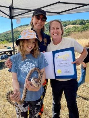 A young girl holds a snake as a woman holds a resolution while another woman stands behind the child
