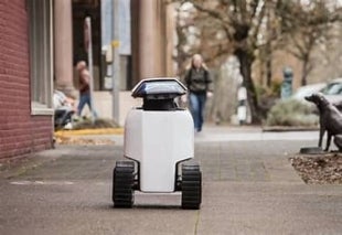 A Daxbot robot rolls along a sidewalk navigating past buildings and a street bench