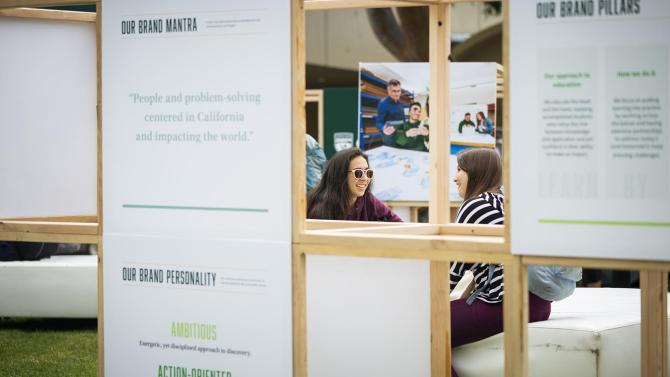 Foreground is a wooden display showing brand information with two students sitting on a cushioned bench in the background.