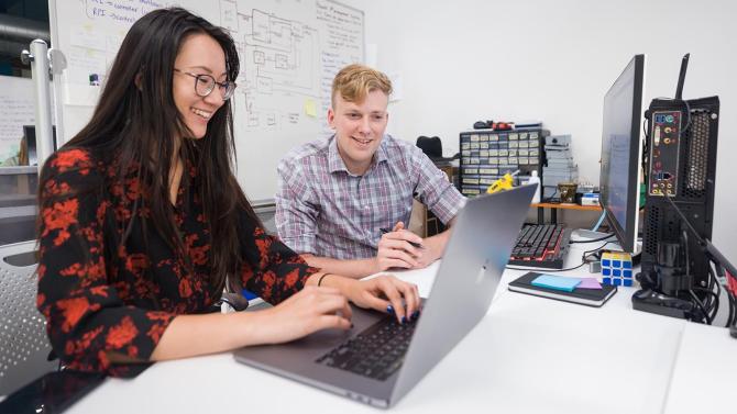 Two students chat while seated at a table with a laptop open in front of them.