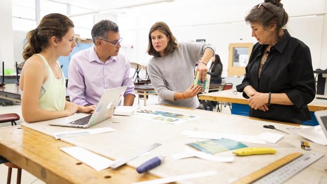 Four people stand and discuss around a table that contains some papers, rulers and a laptop.