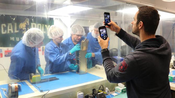 A man holds two phones and takes pictures of students working in the CubeSat cleanroom. 