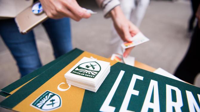 A person takes a sticker of the cal Poly Shield off of a pile of stickers that are stacked atop a Cal Poly pennant. 