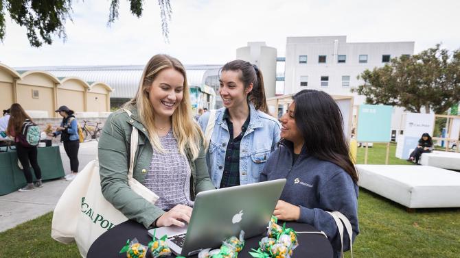 Three students stand talking at a table, looking at a laptop. 