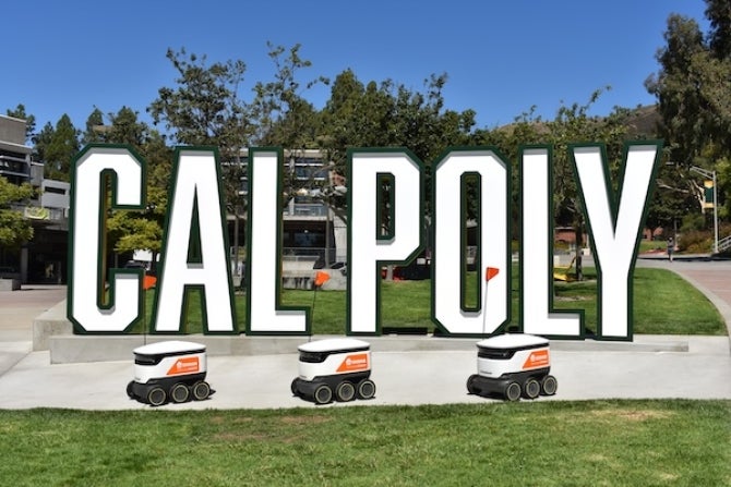 Three Starship robots, which are six-wheeled carriers under 30 inches long, 2 feet wide and tall, with a small orange flag and Grubhub branding, are parked in front of a structure of the letters spelling out Cal Poly in the University Union on campus.