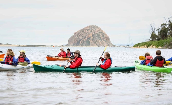 Groups of tudents and their clients paddle kayaks in Morro Bay with Morro Rock in the background