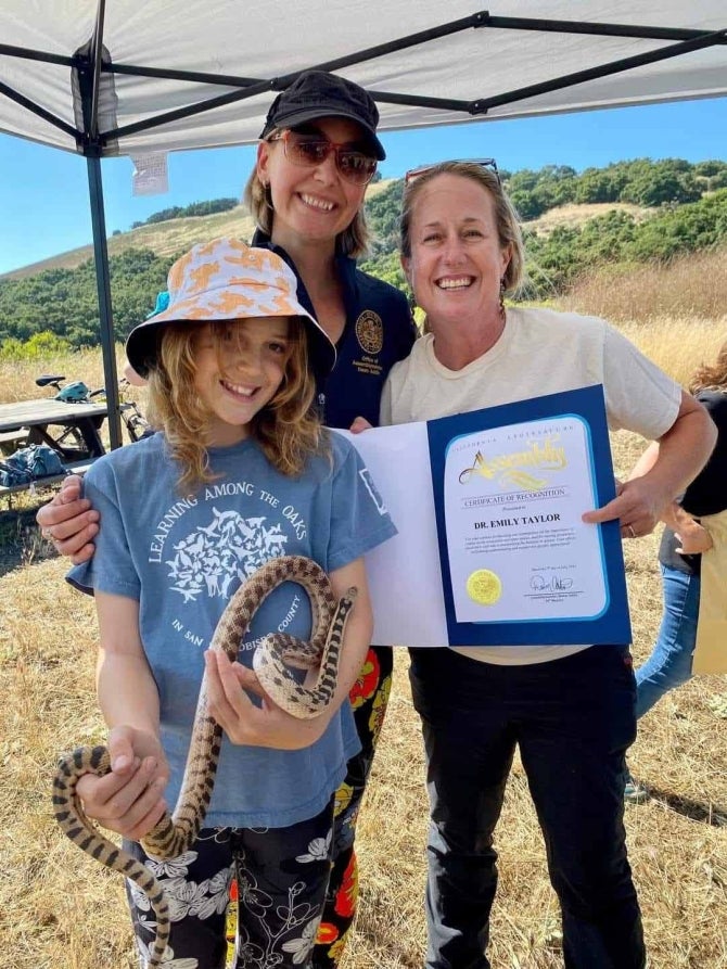 A young girl holds a snake as a woman holds a resolution while another woman stands behind the child