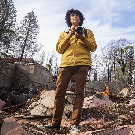 Man holds a camera while standing in the wreckage of the town of Paradise.