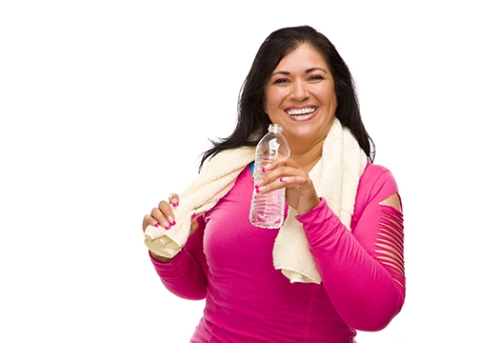 A smiling woman in a pink warmup suit with a towel over her shoulders holds a water bottle