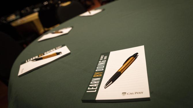 A Cal Poly branded notepad and pen sit at each place setting around a table at an event. 