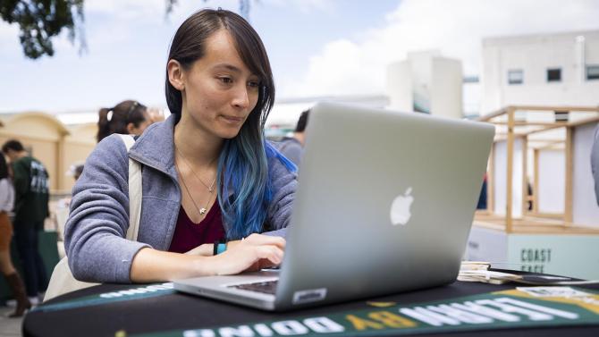 A student stands at a table outside on a laptop.