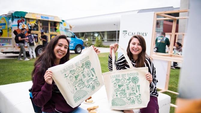 Two students sit on an outdoor bench and hold up screenprinted Cal Poly bags. 