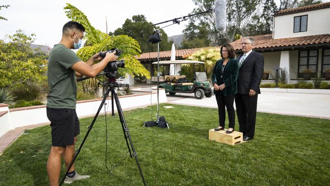 A man adjusts a video camera aimed at Jeff and Sharon Armstrong who are standing in front of their house. 