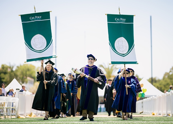 Commencement stage officials holding college banner including a man carrying the ceremonial mace enter the stadium for the 2023 Cal Poly commencement