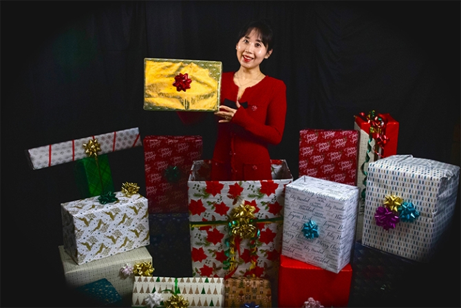 Assistant professor of economics Miranda Yi holds a decorated preseent while standin in a wrapped box surrounded by about a dozen colorfully wrapped packages